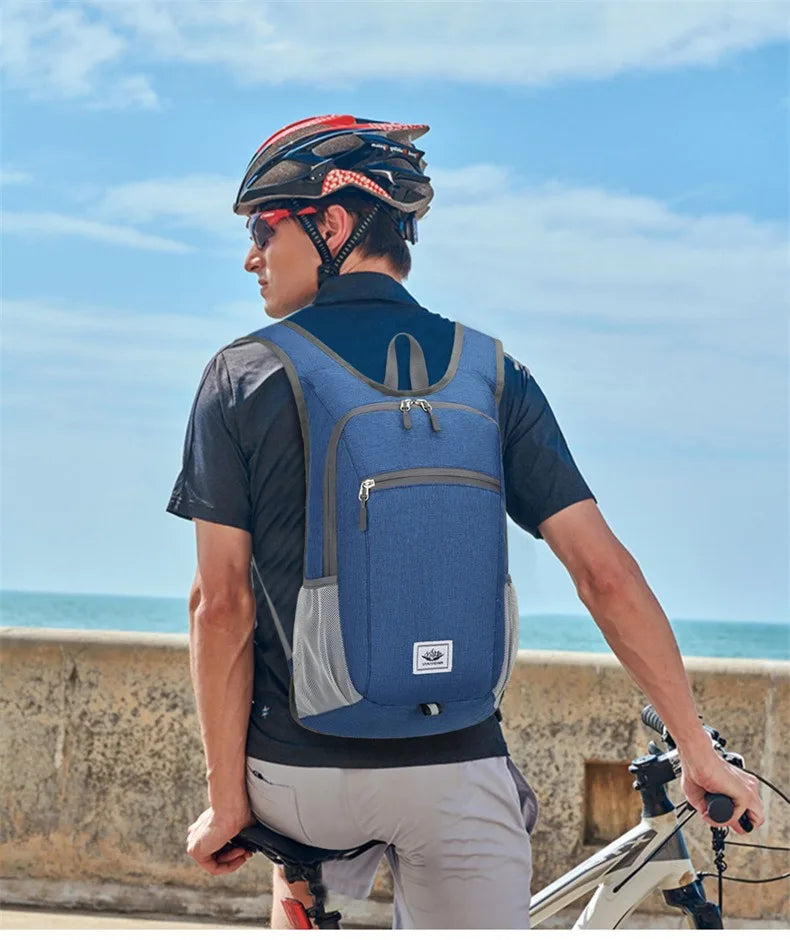 A man wearing a blue and gray cycling backpack, a black cycling helmet, and sunglasses, standing on a beach with a bicycle in the background.