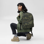 Camouflage backpack worn while kneeling, showing front pocket and adjustable straps.