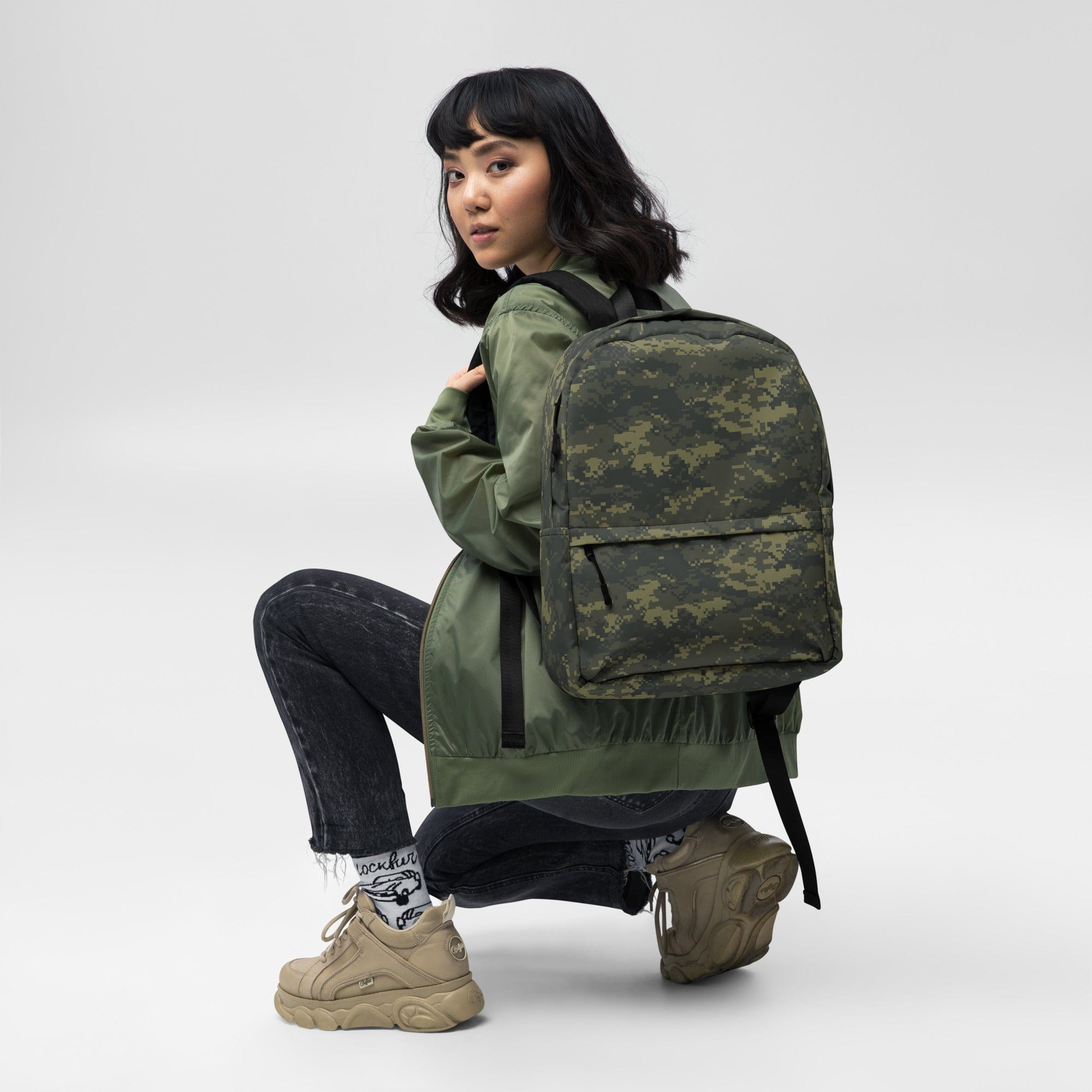 Camouflage backpack worn while kneeling, showing front pocket and adjustable straps.