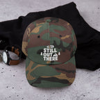 Camo baseball cap with embroidered “STILL OUT THERE” logo on front, curved brim.