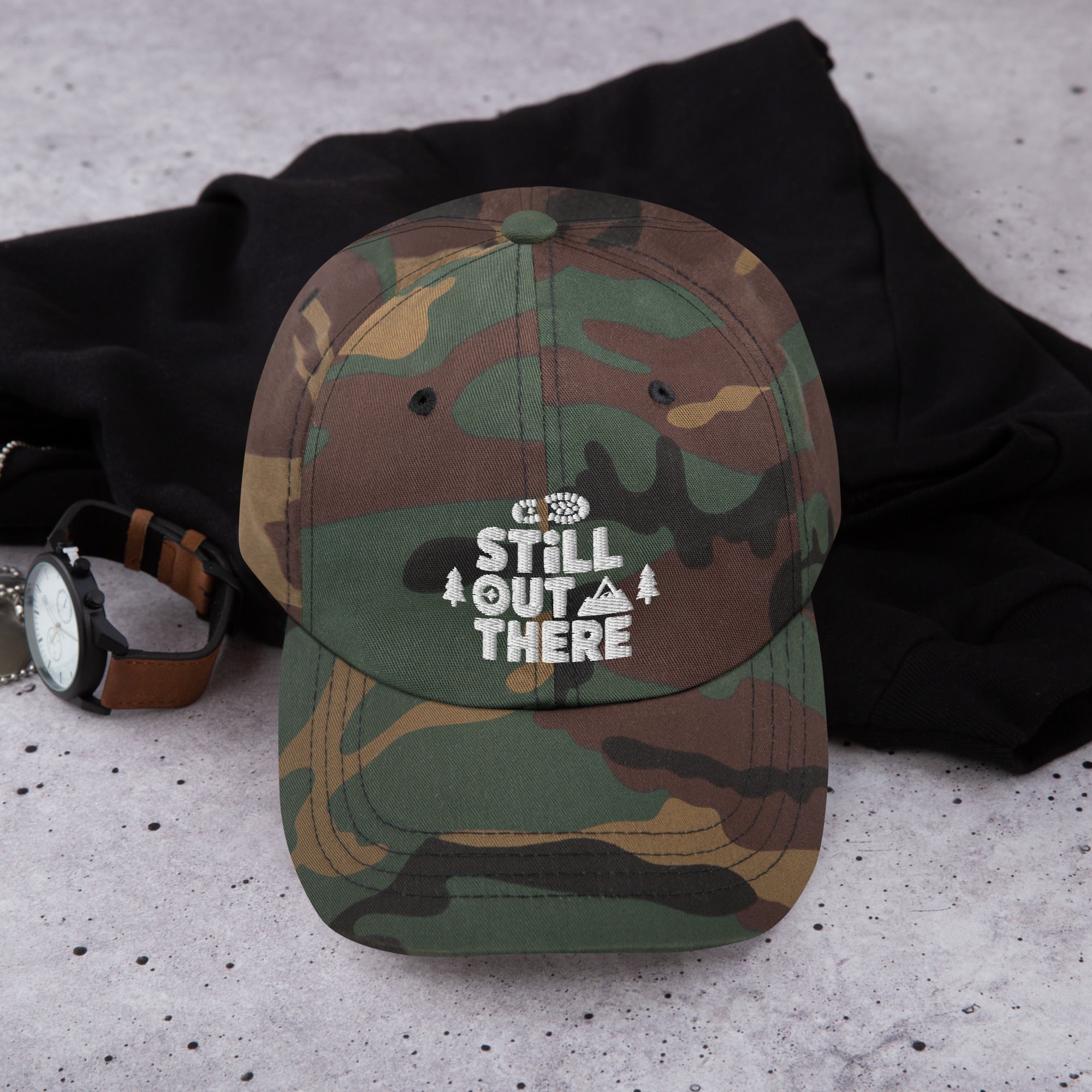 Camo baseball cap with embroidered “STILL OUT THERE” logo on front, curved brim.