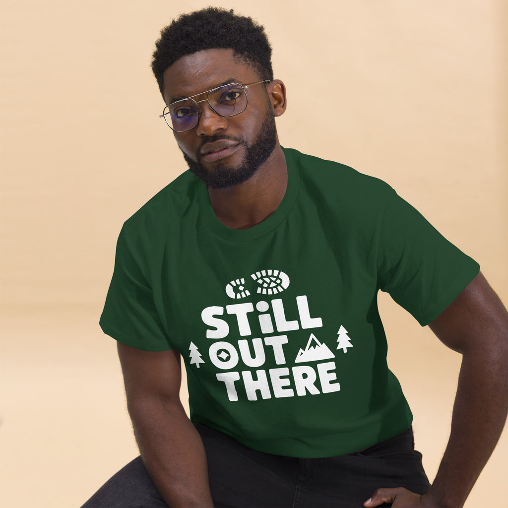 Forest green hiking T-shirt with white “STILL OUT THERE” logo and tree graphics
