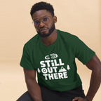 Forest green hiking T-shirt with white “STILL OUT THERE” logo and tree graphics