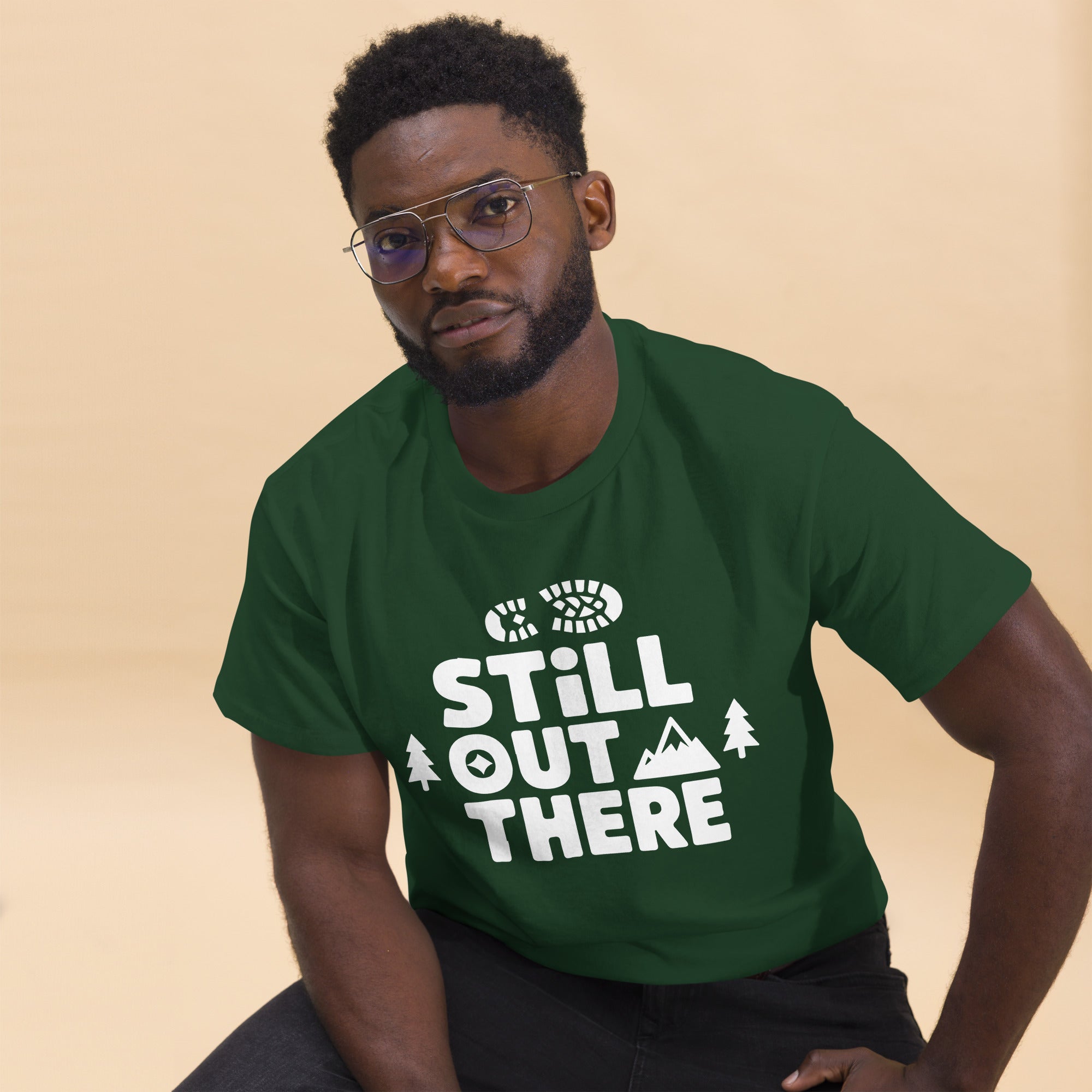 Forest green hiking T-shirt with white “STILL OUT THERE” logo and tree graphics